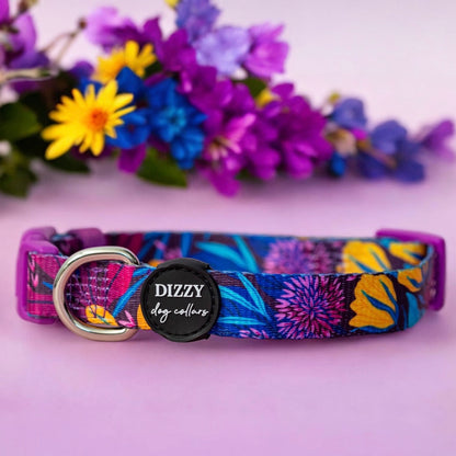 A colourful dog collar with a vibrant floral pattern, featuring a silver D-ring and a black label with "Dizzy Dog Collars" text. The collar is displayed against a soft pink background with a bouquet of purple, blue, and yellow flowers in the background, creating a lively and visually appealing scene.