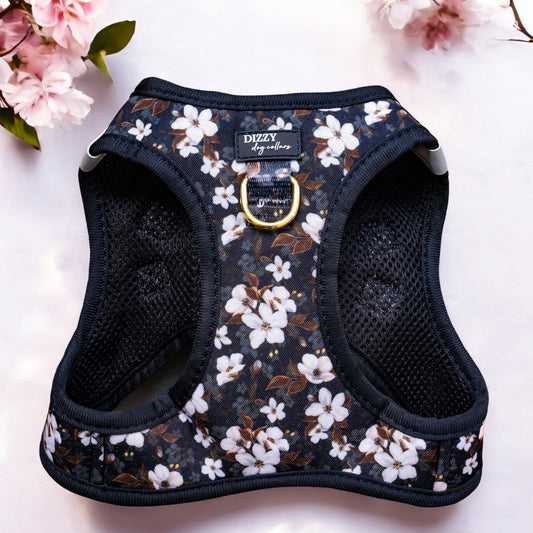 This image showcases a black dog harness with a floral pattern, featuring white flowers with brown and green accents on a dark background. The harness has a breathable mesh design for comfort and a gold D-ring for leash attachment. The brand name "DIZZY dog collars" is visible on a label stitched onto the harness. The harness is placed on a light grey surface, with pink flowers positioned in the top left and right corners of the image, adding a soft, natural touch to the presentation.